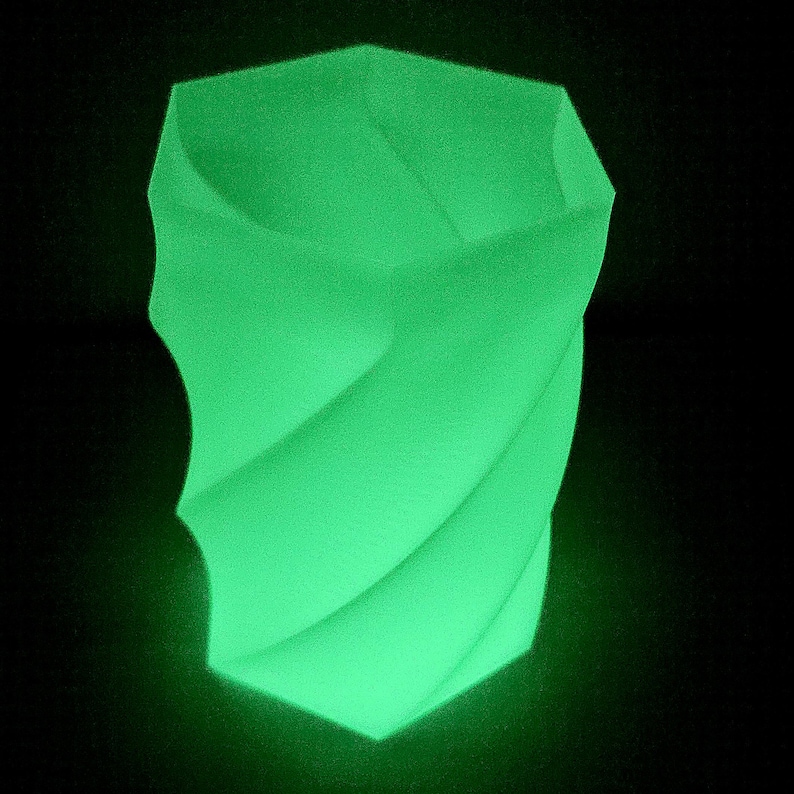 Op de afbeelding: Een gloeiende groene, geometrische vaas met een gedraaid ontwerp. De vaas heeft een unieke vorm met een brede opening en een gestructureerd oppervlak. De heldergroene kleur contrasteert met de zwarte achtergrond en benadrukt de vorm van de vaas.