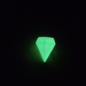 May include: A luminous green diamond-shaped object against a black backdrop. The object glows brightly, creating a vibrant appearance. The diamond shape is well-defined, with sharp angles and a pointed base.