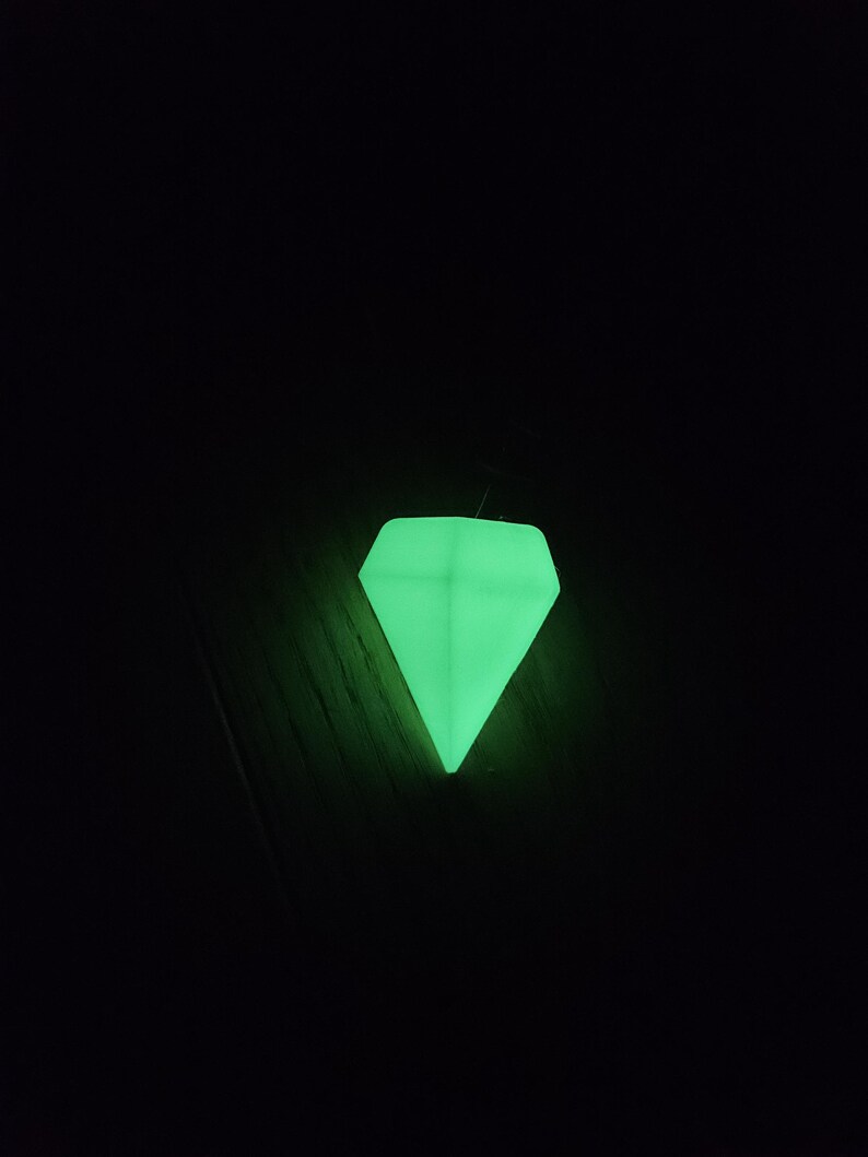 May include: A luminous green diamond-shaped object against a black backdrop. The object glows brightly, creating a vibrant appearance. The diamond shape is well-defined, with sharp angles and a pointed base.