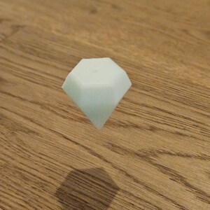May include: A pale green, diamond-shaped object floats above a wooden surface. The object's shadow is visible below. The diamond shape has multiple facets, reflecting light. The object appears to be made of a solid material.