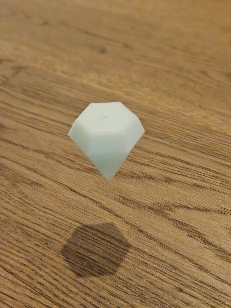 May include: A pale green, diamond-shaped object floats above a wooden surface. The object's shadow is visible below. The diamond shape has multiple facets, reflecting light. The object appears to be made of a solid material.