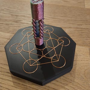 Könnte beinhalten: Ein dekoratives Objekt mit einer zylindrischen, strukturierten, lila und blauen Komponente auf einer schwarzen, geometrischen Basis mit kupferfarbenen Drahtdetails. Die Basis ist sechseckig. Das Objekt wird auf einer Holzoberfläche präsentiert.