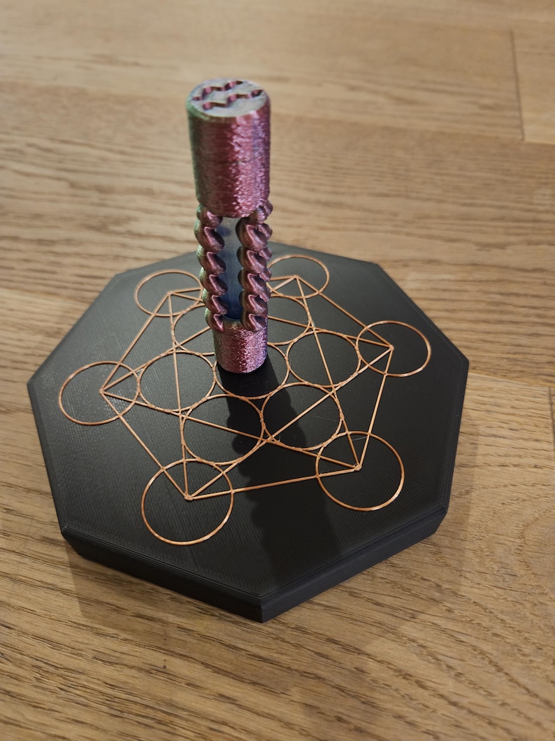 Puede incluir: Un objeto decorativo con un componente cil&iacute;ndrico, texturizado, morado y azul, sobre una base negra geom&eacute;trica con detalles de alambre color cobre. La base es hexagonal. El objeto se muestra sobre una superficie de madera.