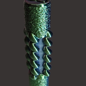 Könnte beinhalten: Zylindrisches, irisierendes grün-blaues Objekt mit strukturierter Oberfläche und einer Reihe von gebogenen, schuppenartigen Vorsprüngen. Die Oberseite weist ein stilisiertes Design auf. Das Objekt ist vertikal vor dunklem Hintergrund ausgerichtet.