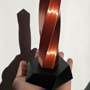K&ouml;nnte beinhalten: Eine kupferfarbene, gedrehte geometrische Skulptur mit strukturierter Oberfl&auml;che, montiert auf einem schwarzen, facettierten Sockel. Der Schatten der Skulptur wird auf einen wei&szlig;en Hintergrund geworfen.