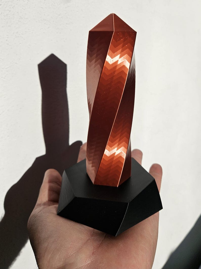 K&ouml;nnte beinhalten: Eine kupferfarbene, gedrehte geometrische Skulptur mit strukturierter Oberfl&auml;che, montiert auf einem schwarzen, facettierten Sockel. Der Schatten der Skulptur wird auf einen wei&szlig;en Hintergrund geworfen.