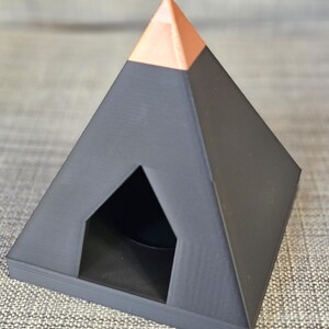 Könnte beinhalten: Ein 3D-gedrucktes, pyramidenförmiges Objekt mit dunkelgrauer Basis und kupferfarbener Spitze. Die Pyramide hat eine dreieckige Öffnung an einer Seite, was auf ein dekoratives oder funktionales Design hindeutet. Das Objekt ist wahrscheinlich aus Kunststoff.