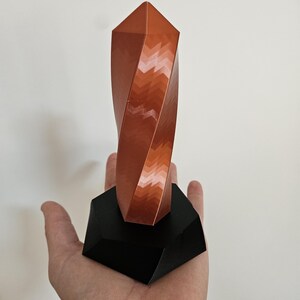 K&ouml;nnte beinhalten: Eine kupferfarbene, geometrische Skulptur mit einem gedrehten Design, montiert auf einem schwarzen, eckigen Sockel. Die Skulptur hat eine facettierte Oberfl&auml;che, die ein Spiel von Licht und Schatten erzeugt. Der Sockel ist dunkel und kontrastreich.
