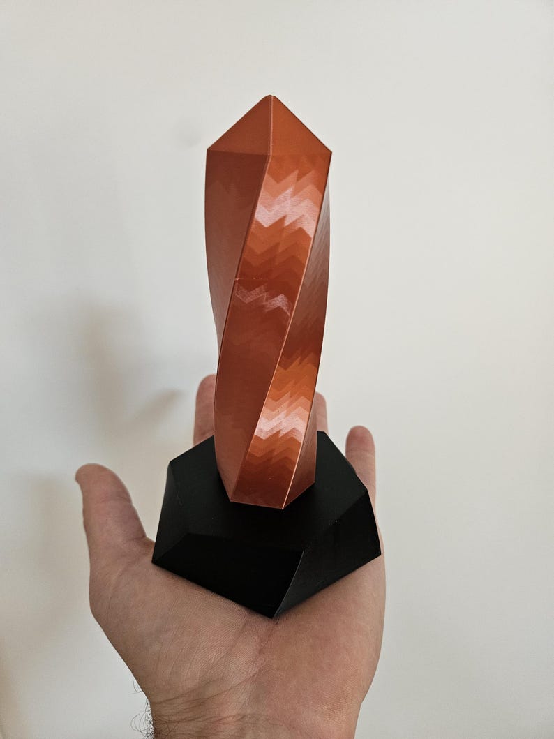 K&ouml;nnte beinhalten: Eine kupferfarbene, geometrische Skulptur mit einem gedrehten Design, montiert auf einem schwarzen, eckigen Sockel. Die Skulptur hat eine facettierte Oberfl&auml;che, die ein Spiel von Licht und Schatten erzeugt. Der Sockel ist dunkel und kontrastreich.