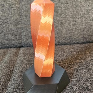 K&ouml;nnte beinhalten: Eine kupferfarbene, geometrische Skulptur mit einem gedrehten Design. Die Skulptur steht auf einem schwarzen, facettierten Sockel. Die Spitze der Skulptur ist spitz. Das St&uuml;ck ist wahrscheinlich 20 cm hoch.