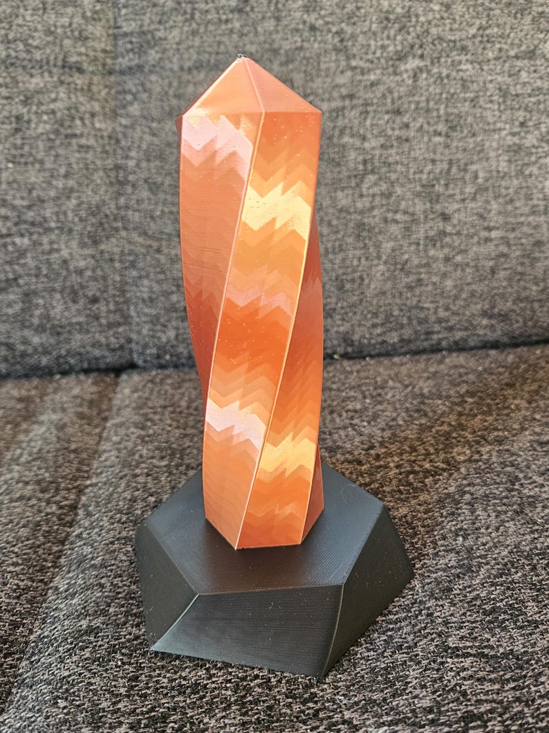 K&ouml;nnte beinhalten: Eine kupferfarbene, geometrische Skulptur mit einem gedrehten Design. Die Skulptur steht auf einem schwarzen, facettierten Sockel. Die Spitze der Skulptur ist spitz. Das St&uuml;ck ist wahrscheinlich 20 cm hoch.