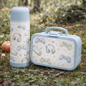 May include: A light blue insulated thermos and lunchbox set. The set features a pattern of video game controllers, headphones, and stars on a white background. The lunchbox has a handle. The thermos and lunchbox are sitting on the ground.