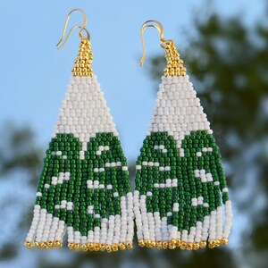 May include: A pair of beaded earrings with a monstera leaf design. The earrings feature a white and green pattern, with gold-colored accents at the top and bottom. The earrings have gold-colored hooks.