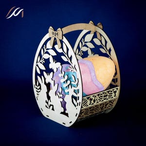 Op de afbeelding: Houten paasmand in de vorm van een ei, met uitgesneden ontwerpen van konijnen, vlinders en bladeren. De mand bevat kleurrijke, eivormige items. De handgreep heeft een strik. De mand is gemaakt van lichtgekleurd hout.