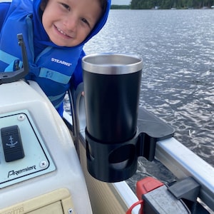 Könnte beinhalten: Ein schwarzer Isolierbecher mit silbernem Rand wird in einem schwarzen Getränkehalter an einem Boot gehalten. Eine junge Person mit einer blauen Kapuzenjacke und Schwimmweste lächelt in die Kamera. Das Boot befindet sich auf einem See.