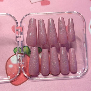 May include: A set of ten pink, glitter-covered, coffin-shaped false nails arranged in a clear plastic case. The nails are a light pink color with a sparkly, iridescent finish. The case is open, revealing the full set.