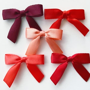 May include: Five satin ribbon bows in various shades of red, from burgundy to coral. The bows are arranged in a semi-circle on a white background.