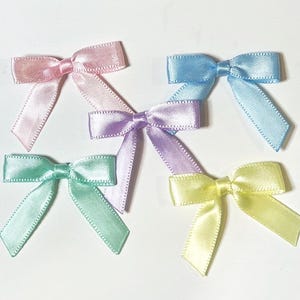 May include: Five small satin bows in pastel colors: pink, blue, purple, green, and yellow. Each bow has a classic bow shape with two loops and two streamers. The bows are arranged on a white surface.