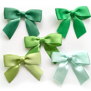 May include: Five satin bows in shades of green and aqua. The bows are arranged on a white background. Each bow has a classic bow shape with two loops and two streamers. The colors range from a deep forest green to a light aqua.