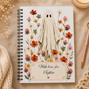 May include: A spiral-bound notebook featuring a watercolor illustration of a white ghost surrounded by colorful wildflowers. A rectangular label at the bottom contains the text "With love, for Sophia" with a small red heart icon below the name.