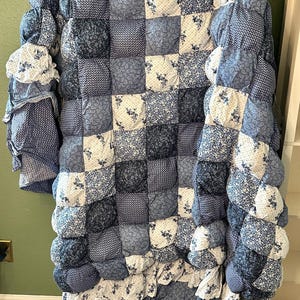 May include: A blue and white patchwork quilt featuring a puff-style design with various floral, paisley, and geometric patterns. The quilt is constructed from individual square fabric sections and includes a ruffled edge along the bottom border.
