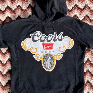 May include: Black hooded sweatshirt with a Coors Banquet Beer logo patch on the back. The patch is white with red and yellow accents.