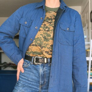 May include: A blue and black checkered flannel shirt with a camouflage t-shirt underneath. The shirt is buttoned up and has two pockets on the front. The person is wearing blue jeans and a silver belt.