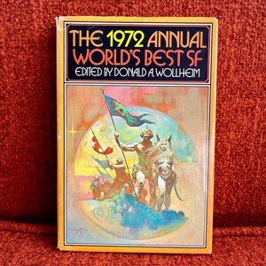 Puede incluir: La portada del libro de antología de ciencia ficción "The 1972 Annual World's Best SF", editado por Donald A. Wollheim, con una ilustración colorida de una escena de fantasía con un guerrero, una mujer y una bandera.