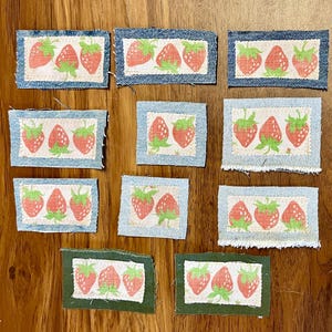 May include: A collection of ten fabric patches featuring a strawberry design. The strawberries are red with green stems and leaves, set against a light blue background. The patches are rectangular, with a denim-like border.