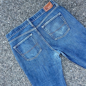 May include: A pair of blue denim jeans with a brown leather patch on the back waistband. The jeans have two back pockets with red stitching.