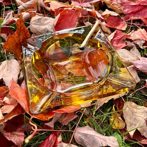 May include: A clear glass ashtray with a yellow tint sits on a bed of fallen autumn leaves. The ashtray has a rounded rectangular shape with a slight lip around the edge. A single cigarette is resting inside the ashtray.