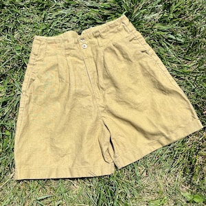 Puede incluir: Pantalones cortos color caqui con cintura alta y pliegues. Los pantalones cortos tienen dos botones en la cintura y bolsillos laterales. Los pantalones cortos están hechos de una tela resistente y están sobre hierba verde.