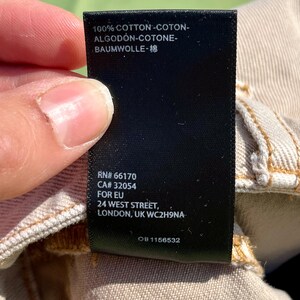 Puede incluir: Una etiqueta de ropa negra con texto blanco, sostenida por una persona. La etiqueta dice "100% COTTON" en varios idiomas, un c&oacute;digo de producto y una direcci&oacute;n en Londres.