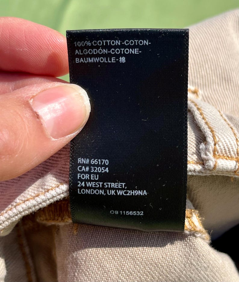 Puede incluir: Una etiqueta de ropa negra con texto blanco, sostenida por una persona. La etiqueta dice "100% COTTON" en varios idiomas, un c&oacute;digo de producto y una direcci&oacute;n en Londres.