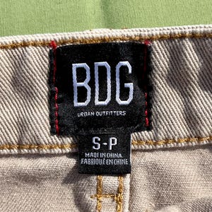 Puede incluir: Primer plano de una tela beige con una etiqueta negra. La etiqueta presenta las letras blancas "BDG" sobre "URBAN OUTFITTERS". Debajo, se lee "S-P" y "MADE IN CHINA. FABRIQU&Eacute; EN CHINE".
