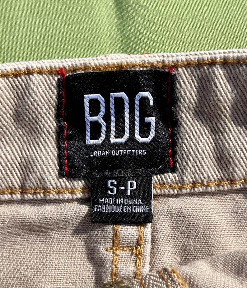 Puede incluir: Primer plano de una tela beige con una etiqueta negra. La etiqueta presenta las letras blancas "BDG" sobre "URBAN OUTFITTERS". Debajo, se lee "S-P" y "MADE IN CHINA. FABRIQU&Eacute; EN CHINE".