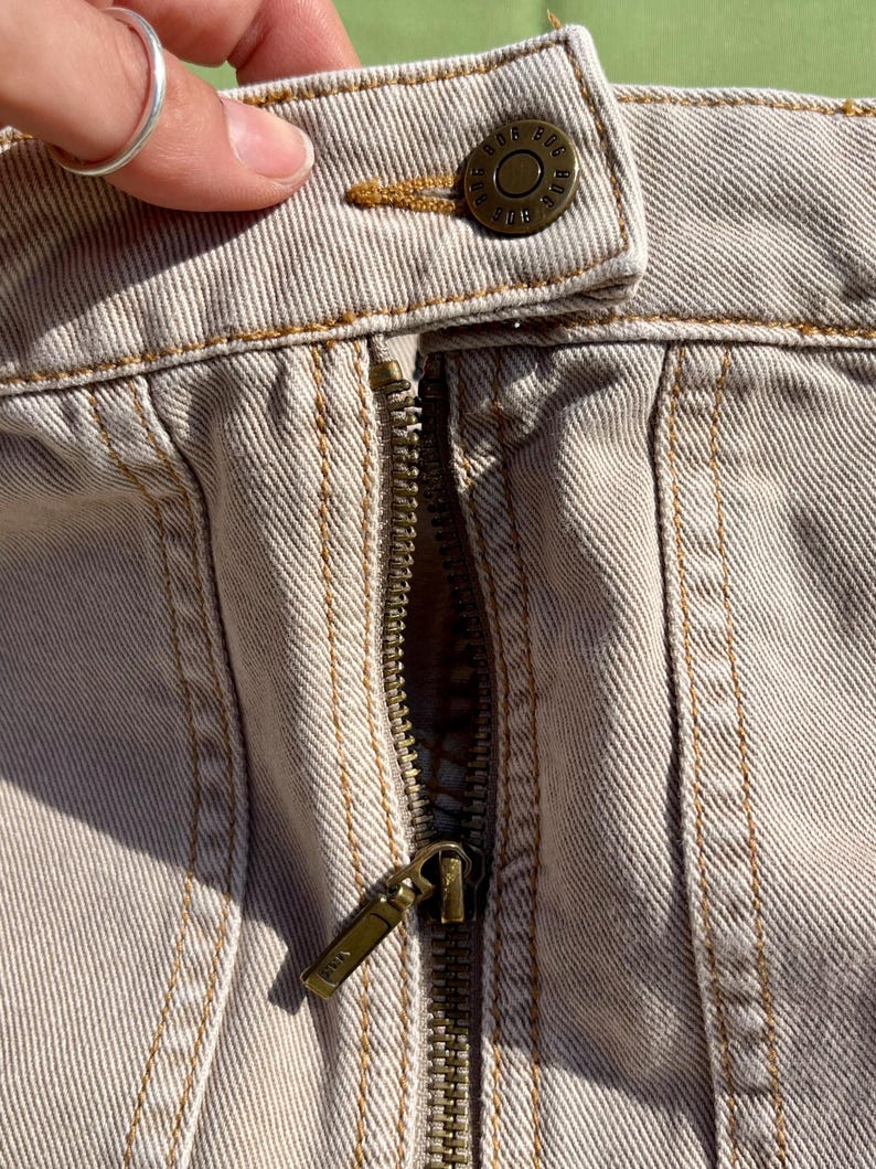 Puede incluir: Primer plano de tejido vaquero beige con cremallera y cierre de bot&oacute;n de lat&oacute;n. El bot&oacute;n est&aacute; grabado con el texto "BDG". Los detalles de costura visibles y el tirador de cremallera met&aacute;lico a&ntilde;aden textura a la prenda.