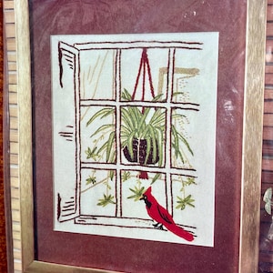 May include: A framed cross-stitch embroidery depicting a window with a hanging plant and a red cardinal bird. The embroidery is on a white background with brown stitching. The frame is gold with a red mat.