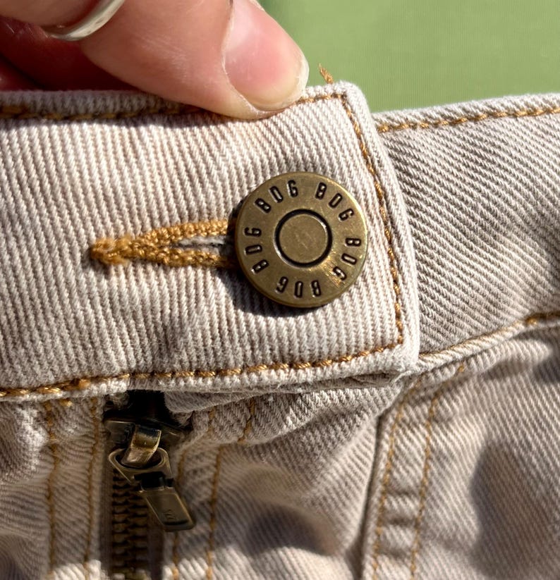 Puede incluir: Primer plano de una prenda vaquera beige con un bot&oacute;n y cremallera de lat&oacute;n. El bot&oacute;n est&aacute; grabado con el texto "806 908 BDC". La costura es de color marr&oacute;n claro en contraste.