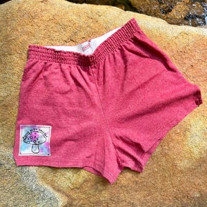 Peut inclure: Short de sport rouge avec un patch tie-dye représentant un champignon et le texte "Ghost Pipe Delights".