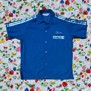 以下が含まれることがあります： 白い縁取りと青いストライプが袖についた青い半袖シャツ。シャツはボタン留めになっており、左側に青と白のチェック柄のポケットがあり、白い糸で「Steve」と刺繍されています。