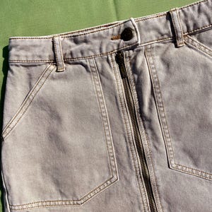 Puede incluir: Primer plano de una falda vaquera gris claro con cremallera frontal y dos bolsillos delanteros. La falda tiene costuras marrones y un cierre de bot&oacute;n en la cintura. La falda est&aacute; hecha de un material vaquero resistente.