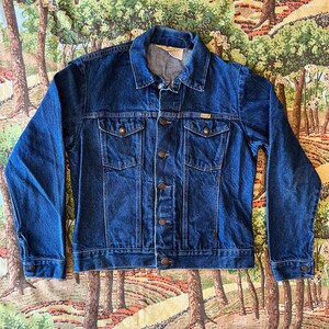 May include: A vintage blue denim jacket with a button closure, two chest pockets, and a Wrangler label on the pocket.