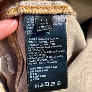 Puede incluir: Primer plano de una etiqueta de ropa negra con texto blanco. La etiqueta incluye instrucciones de lavado en ingl&eacute;s y franc&eacute;s, junto con informaci&oacute;n de tallas: US S, UK S y 170/66A. La etiqueta est&aacute; adherida a una tela beige.