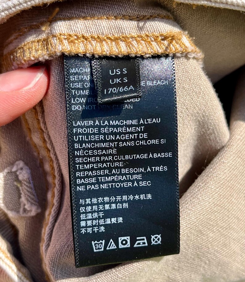 Puede incluir: Primer plano de una etiqueta de ropa negra con texto blanco. La etiqueta incluye instrucciones de lavado en ingl&eacute;s y franc&eacute;s, junto con informaci&oacute;n de tallas: US S, UK S y 170/66A. La etiqueta est&aacute; adherida a una tela beige.