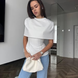May include: A white, short-sleeved top with a cape-like design, paired with light blue jeans. The top has a fitted waist and a round neckline. A cream-colored clutch is held in the hands. A wristwatch with a silver band is visible.