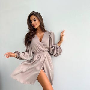 Peut inclure: Robe portefeuille taupe clair avec manches longues et fente haute. La robe présente un décolleté en V et une taille cintrée. Les manches ont un design bouffant au niveau des poignets. La robe est en matière soyeuse.