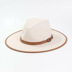 Puede incluir: Un sombrero fedora color crema con una banda y ribete de cuero marrón. El sombrero presenta una copa estructurada y un ala ancha. La banda de cuero tiene una hebilla metálica. El sombrero está sobre un fondo blanco.