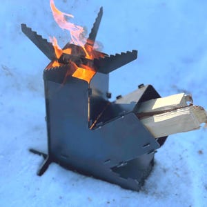 Peut inclure: Un réchaud de camping en métal noir avec un feu à l'intérieur. Le réchaud a un corps rectangulaire avec une ouverture triangulaire pour le bois et un dessus avec des bords dentelés. Les flammes sont orange et jaune.