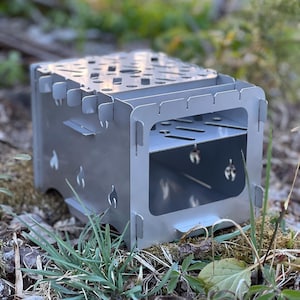 May include: A silver-colored, portable camping stove with a rectangular design. The stove features cut-out patterns on the top and sides, with a shelf inside. It is set on a bed of grass and leaves, suggesting outdoor use.